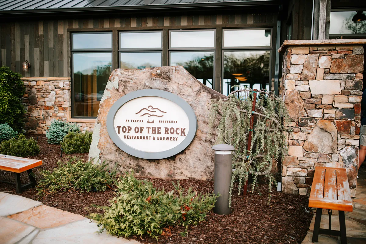 An inviting exterior shot of a restaurant or brewery with stone walls, visible restaurant signage, landscaped greenery, and wooden benches. The outdoor space is designed for dining, socializing, and enjoying craft beer against a scenic backdrop.