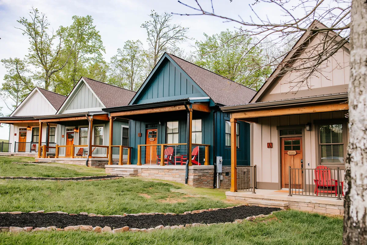 This exterior scene shows several colorful cabins, each with their own porches furnished with rocking chairs, nestled along landscaped pathways. The mood is cozy and social, with people relaxing, chatting, and soaking up the natural surroundings. The setting hints at a community atmosphere while still highlighting the comfort of each individual porch.