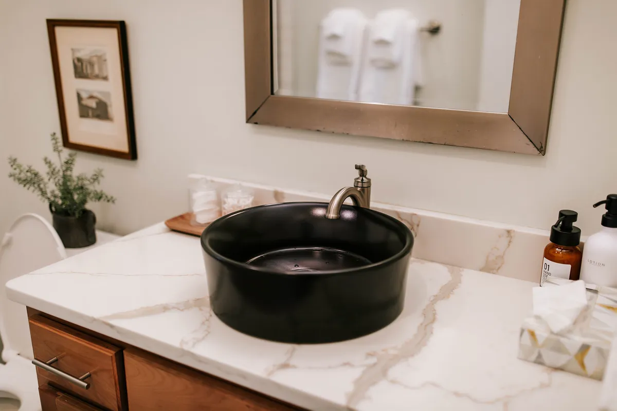 A modern bathroom featuring a sleek black basin sink atop a marble countertop, with a framed mirror and artwork adding decorative touches. The setting is serene and designed for personal relaxation and rejuvenation.
