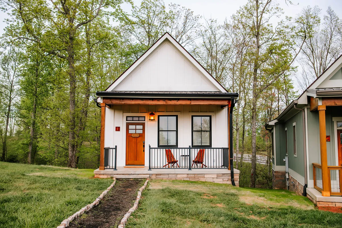This exterior image showcases a welcoming front porch with two classic rocking chairs facing out to the scenic mountain landscape. A sturdy wooden door with elegant glass accents invites guests inside, while warm porch lighting hints at cozy evenings. The setting sun casts a golden glow, suggesting peaceful moments and relaxation in the crisp mountain air.