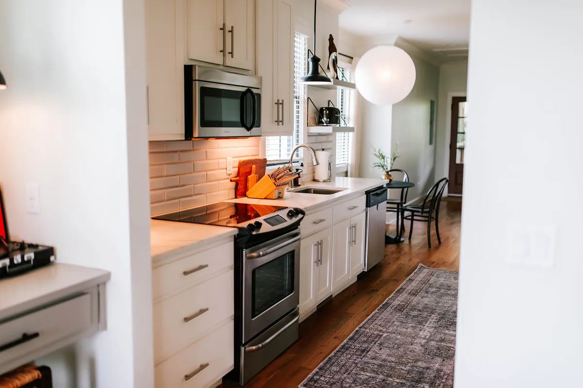 This image presents a well-lit kitchen area featuring a spacious kitchen island, stainless steel appliances, a stylish hanging light fixture, a wooden cutting board, and decorative shelves. The space feels modern, warm, and ready for both everyday meal prep and social cooking experiences.