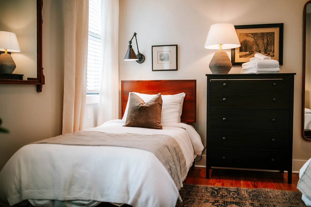 A cozy bedroom with a neatly made bed adorned with pillows, a dresser, a table lamp, and framed artwork. The overall atmosphere is peaceful and stylish, highlighting thoughtful design and comfort.