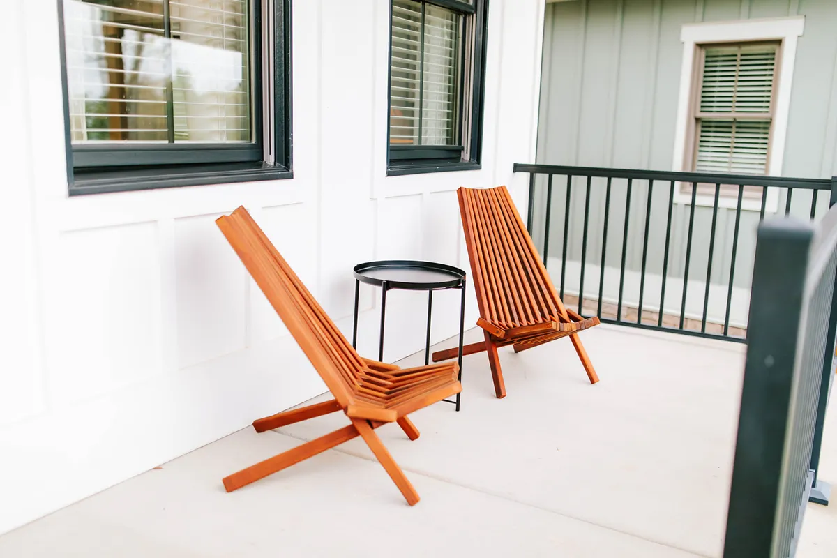 This exterior image captures a covered porch outfitted with wooden chairs and a small black table, situated just outside the cottage's windows. The scene shows an inviting spot perfect for sipping coffee or reading a book, with tranquil views extending out to nature and the mountains beyond.
