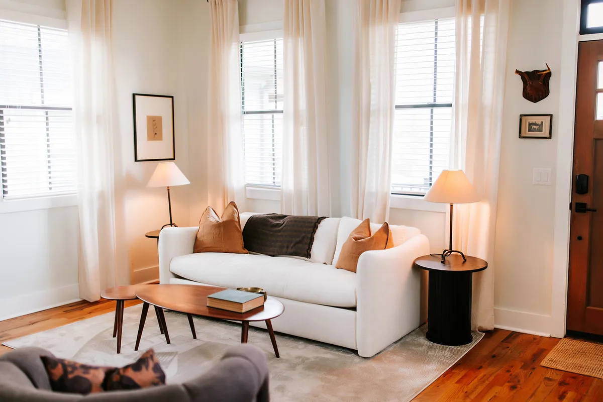 A cozy, well-lit living room with a sofa, coffee table, lamps, and flowing curtains. Natural light streams in through large windows, illuminating the inviting seating area and creating a space ideal for both relaxation and lively conversation.